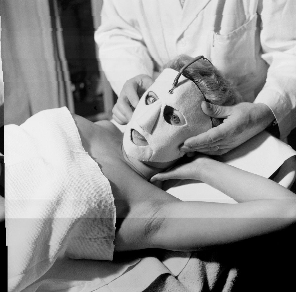 Person lying down with a treatment mask, attended by someone in a white coat, suggesting a skincare or spa setting