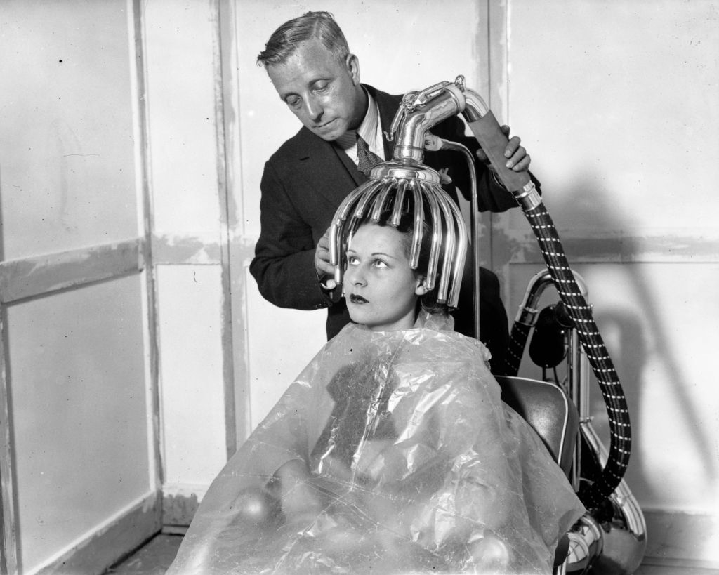 A woman sits under an early 20th-century hair drying machine, with a man adjusting it. She is covered with a plastic cape