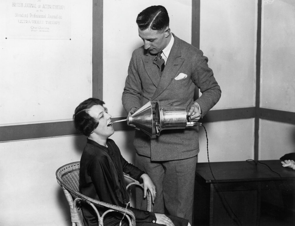 A woman in seated position undergoes a vintage beauty treatment with a man using a retro machine aimed at her face in a clinic setting