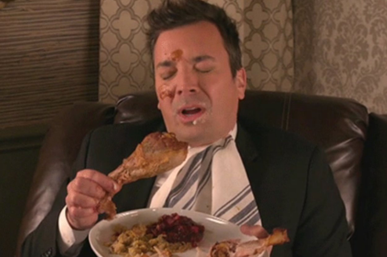 Jimmy Fallon sits on a couch with mashed potatoes on their face, holding a plate with a turkey leg, stuffing, and cranberries, looking full