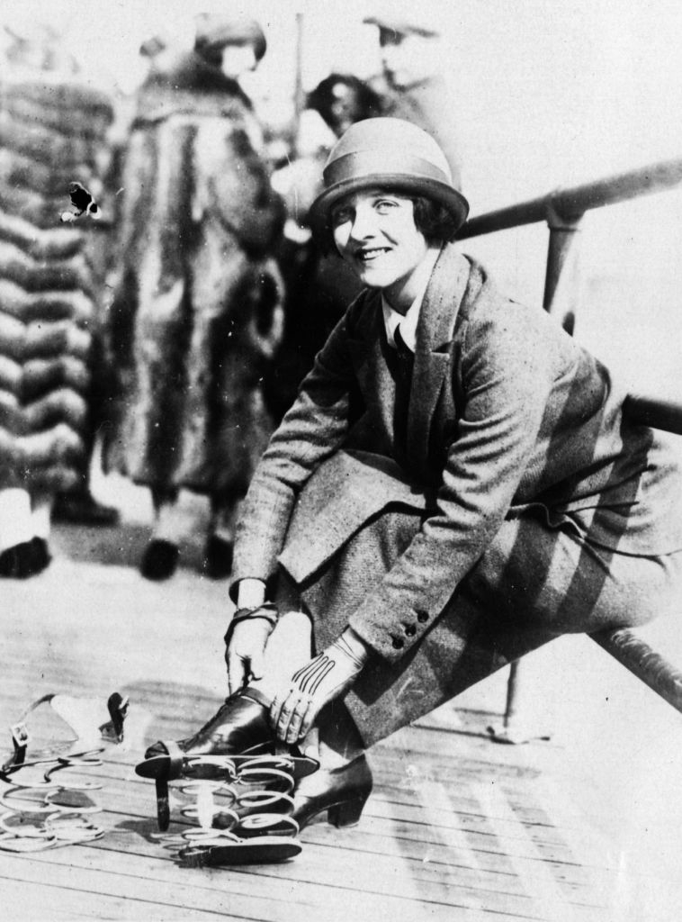 A woman in vintage attire kneels on a deck, adjusting skates attached to her shoes. People with fur coats are blurred in the background