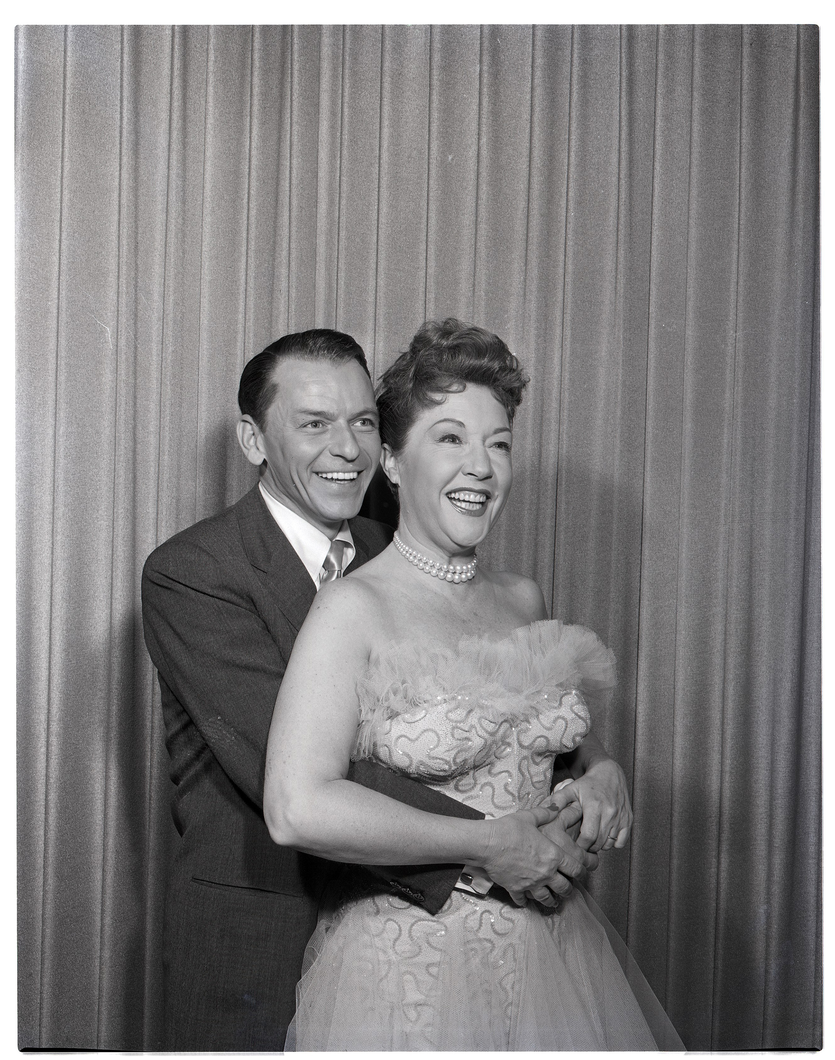 Two people smiling, with one wearing a tuxedo and the other in an elegant, strapless gown with a tulle bodice and patterned skirt, standing in front of curtains