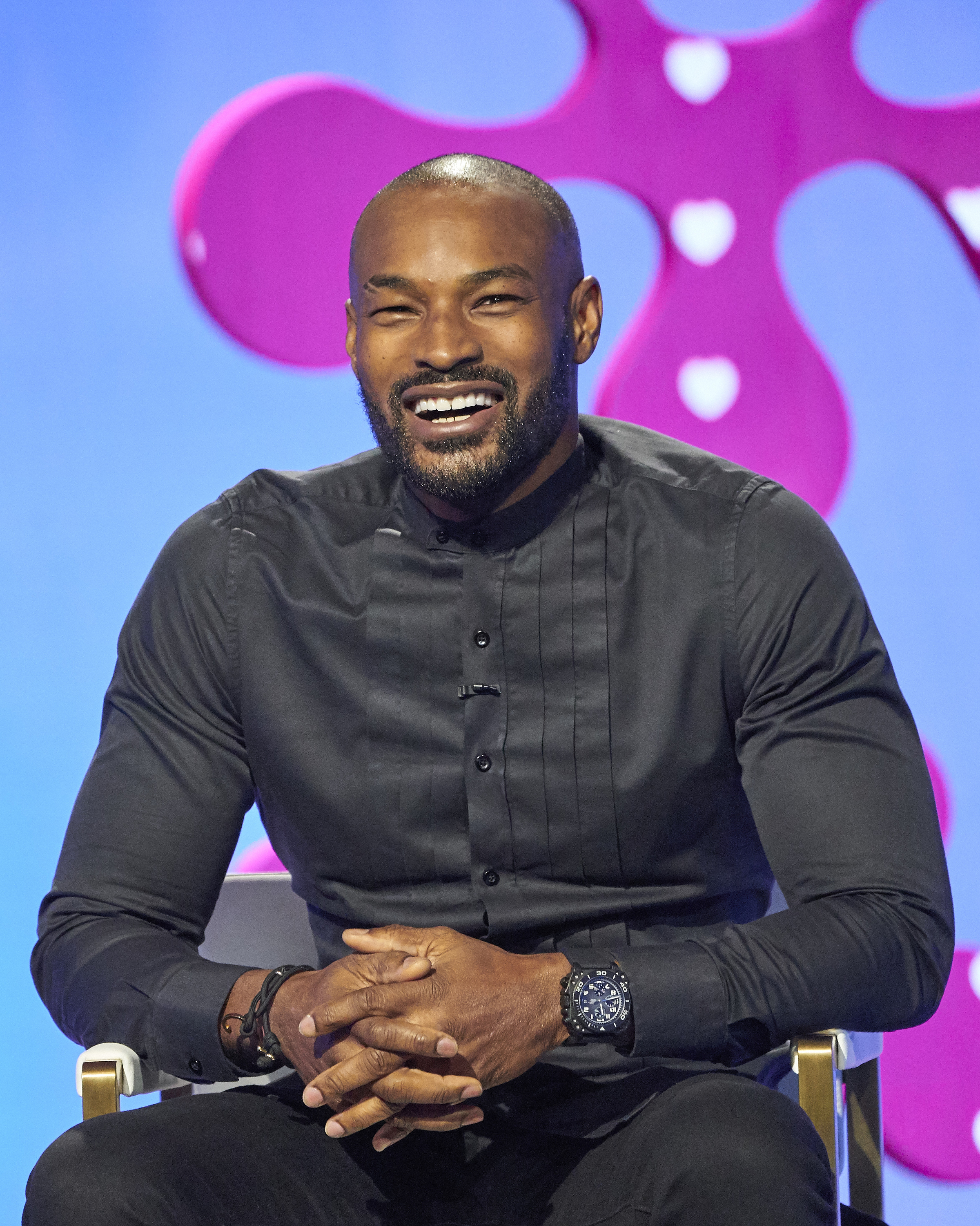 Person smiling, seated, wearing a stylish, fitted long-sleeve shirt with a subtle design. Background features abstract shapes