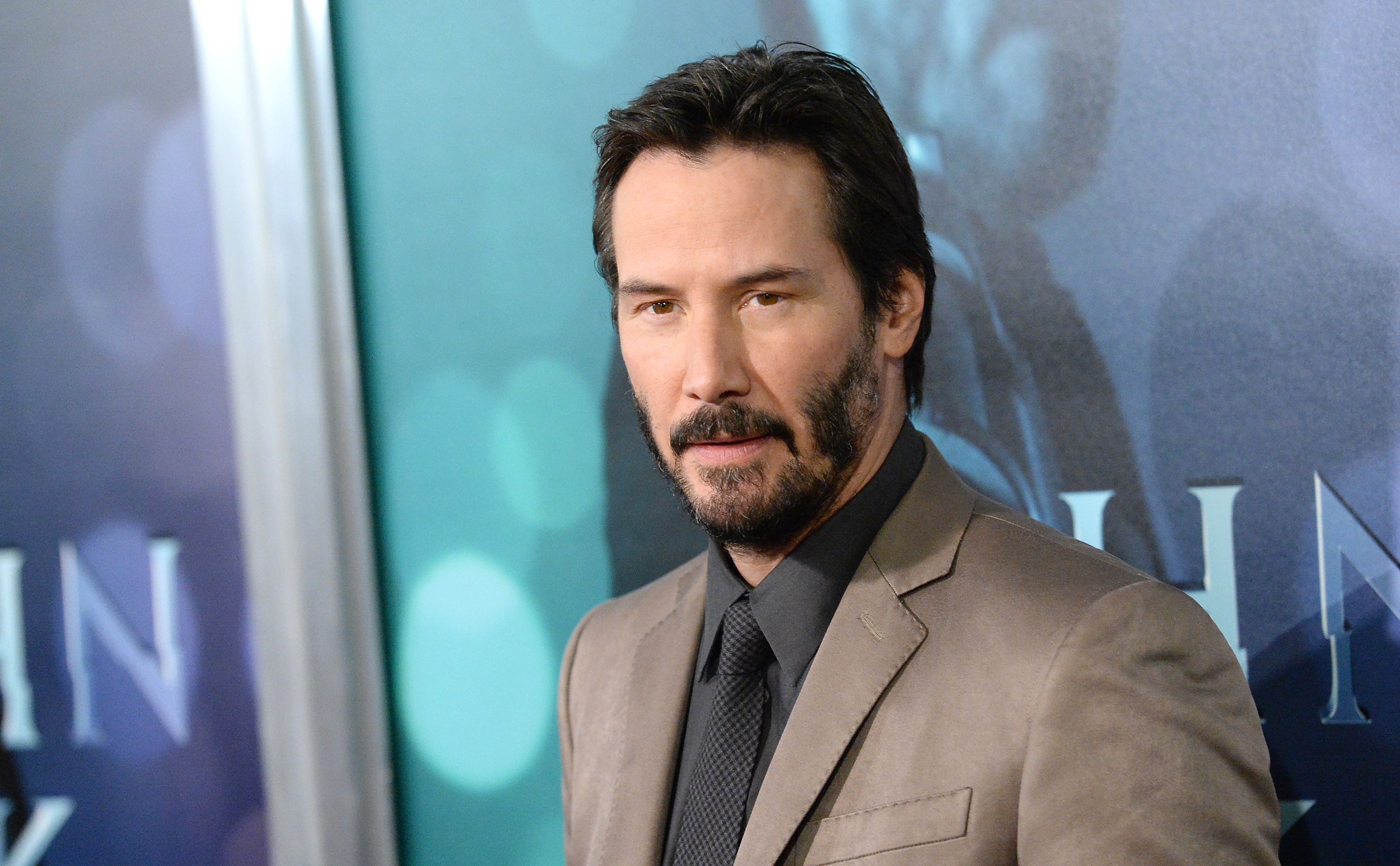 Man in a suit at a movie premiere, standing in front of a backdrop with blurred lights and text