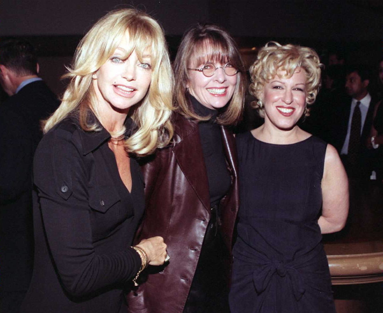 Three women smile at an event. The woman on the left wears a stylish jacket, the center woman wears glasses and a leather coat, and the right woman is in an elegant dress