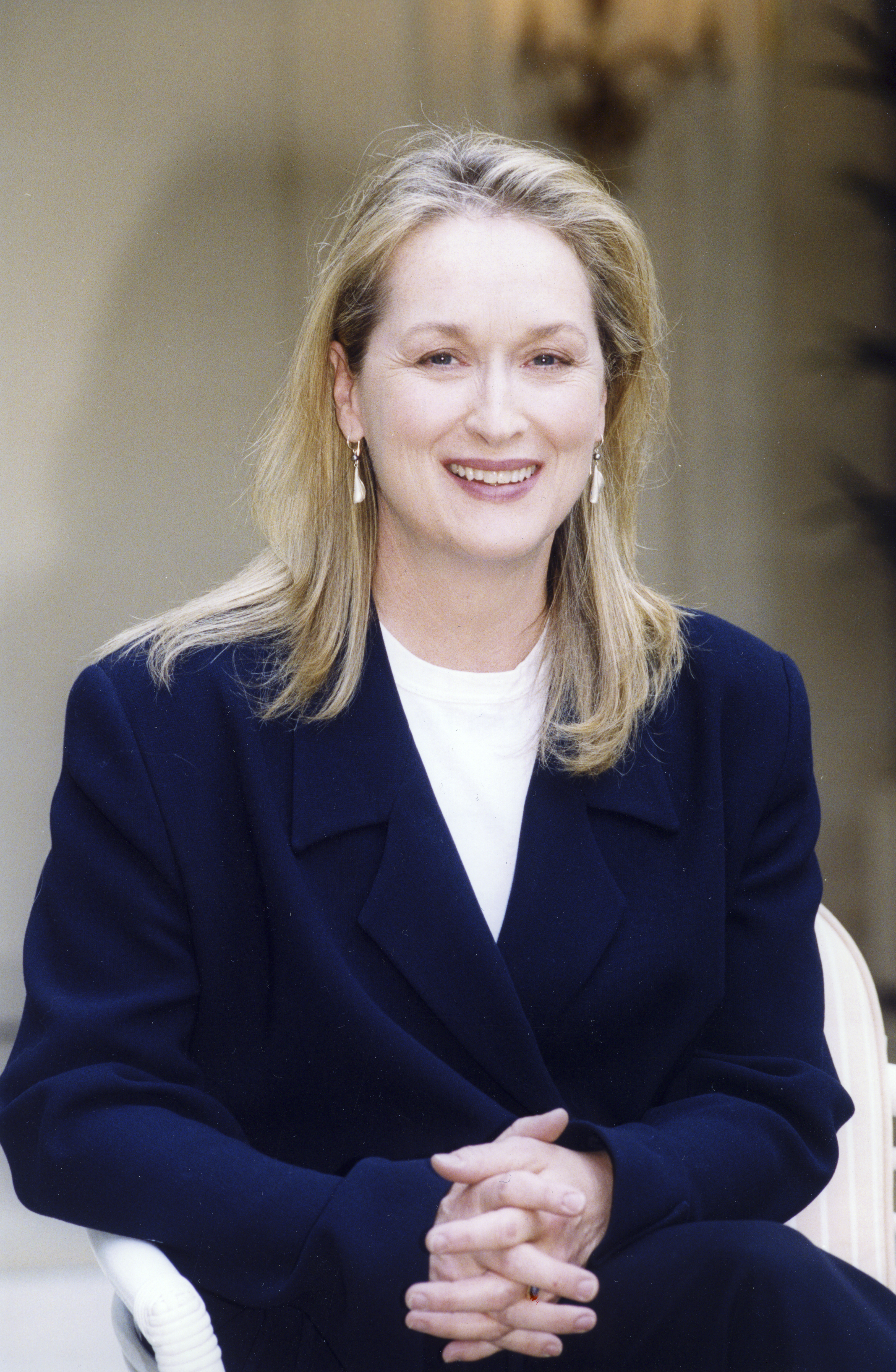 Person smiling, wearing a dark blazer and light shirt, seated with hands clasped