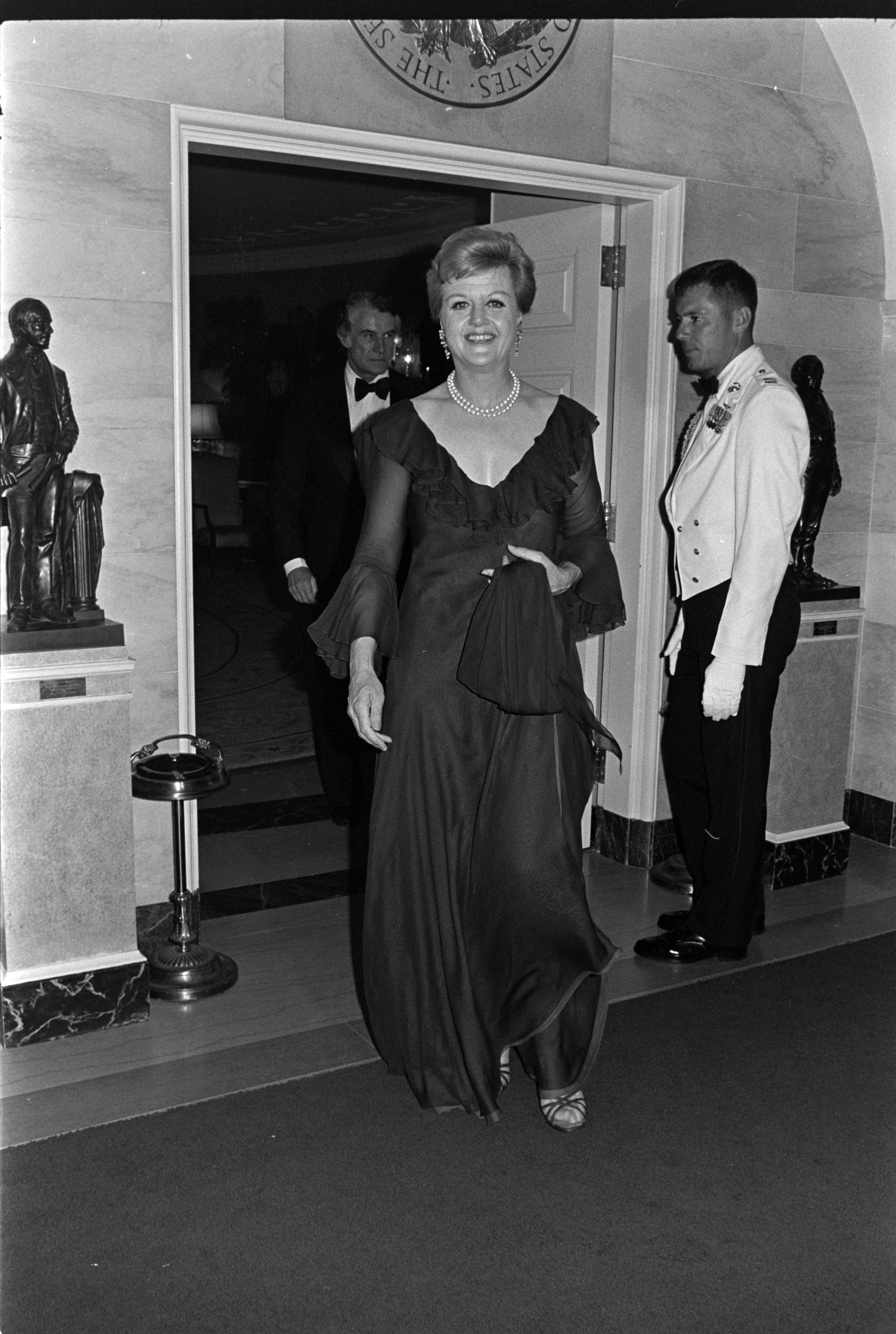 A person in elegant evening attire arrives at a formal event, surrounded by attendees in tuxedos and formal wear