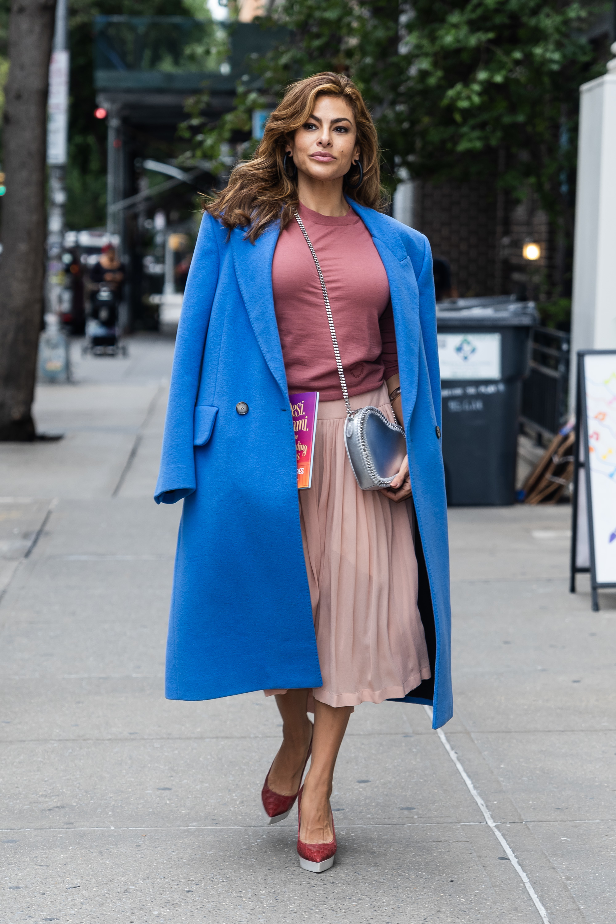 Person walking on a city street, wearing a stylish blue coat, pink pleated skirt, and carrying a crossbody bag