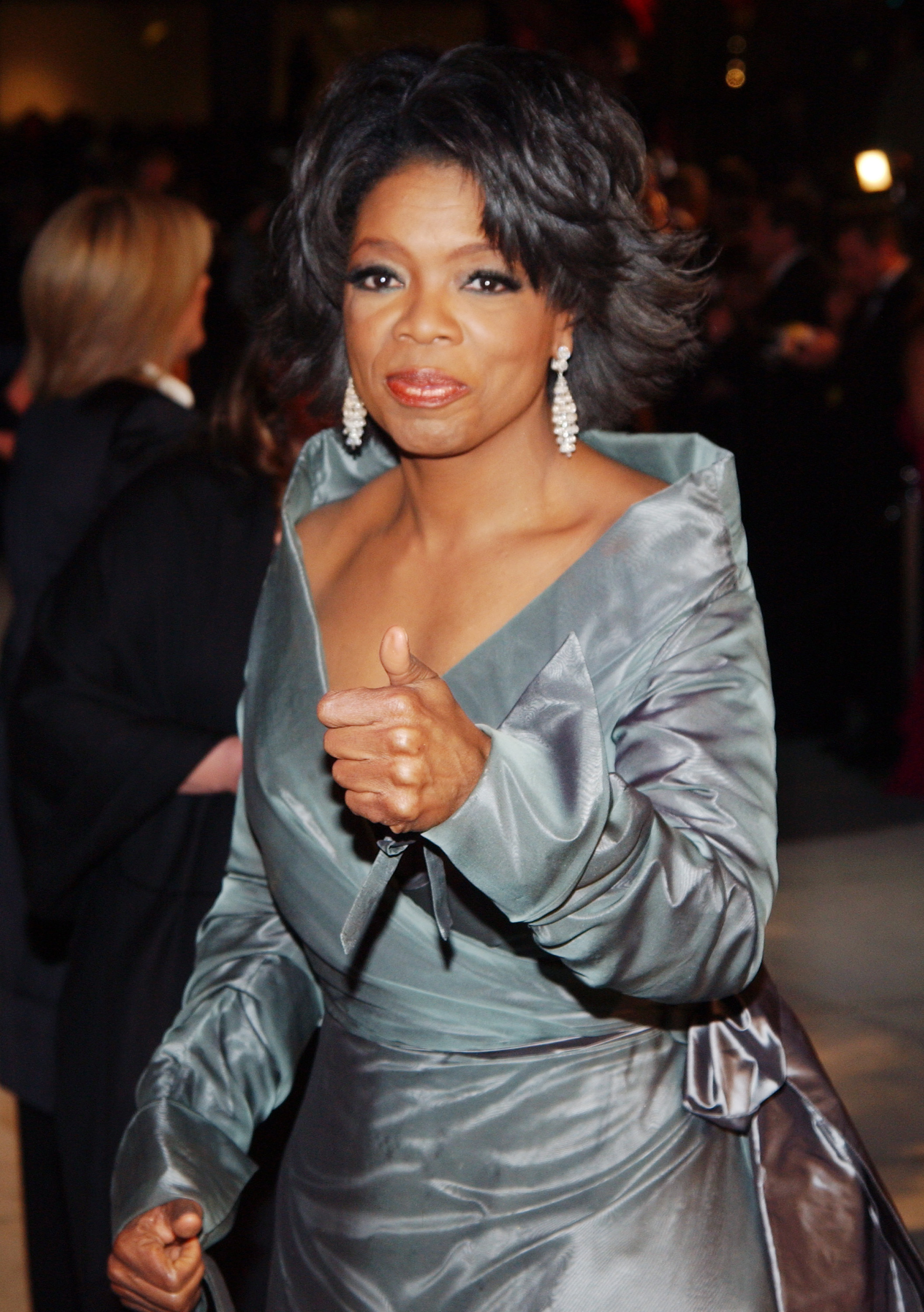 Person in elegant gown on red carpet, giving a thumbs up, wearing chandelier earrings