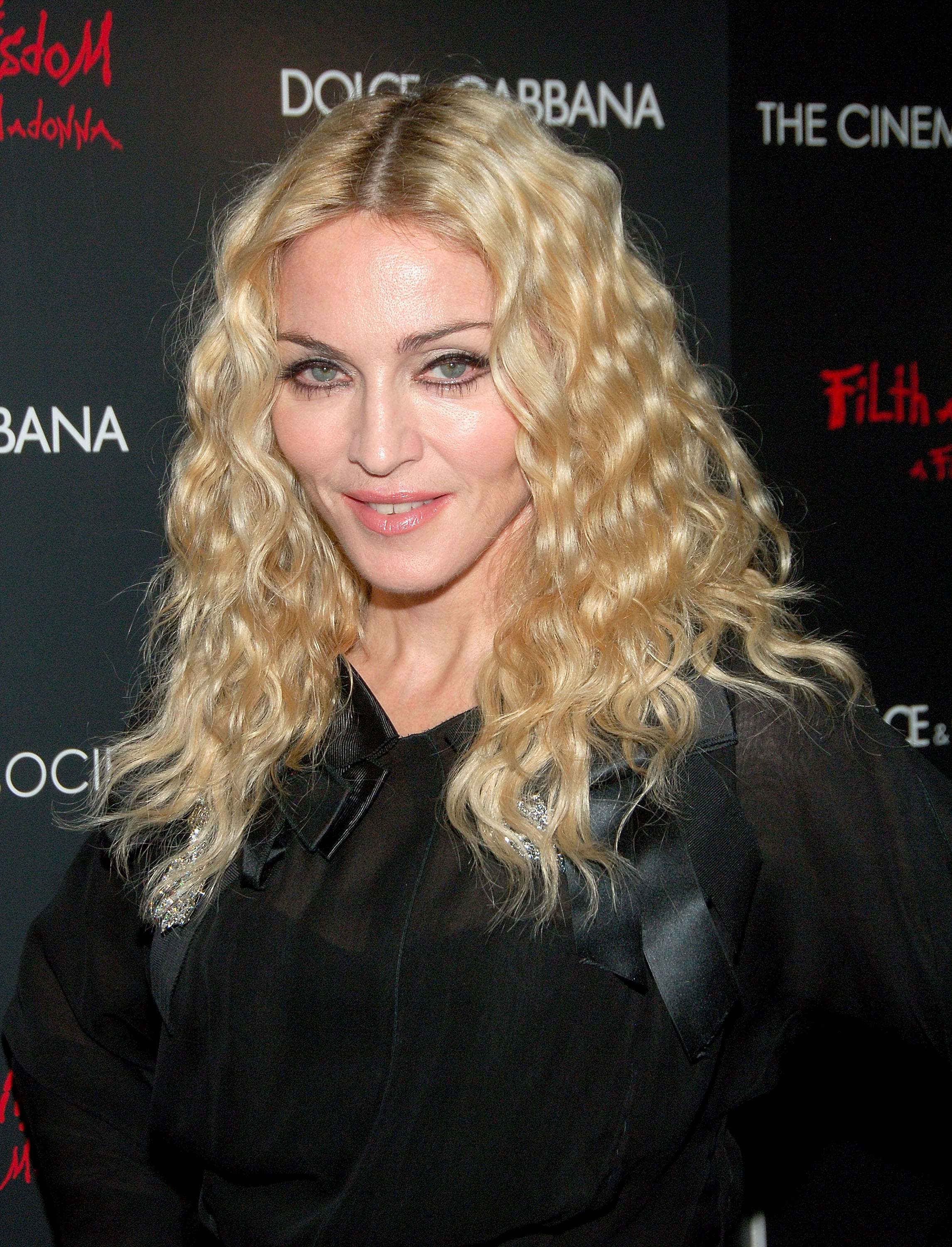 A person on the red carpet in a sheer black top with wavy blonde hair, posing in front of a sponsor backdrop