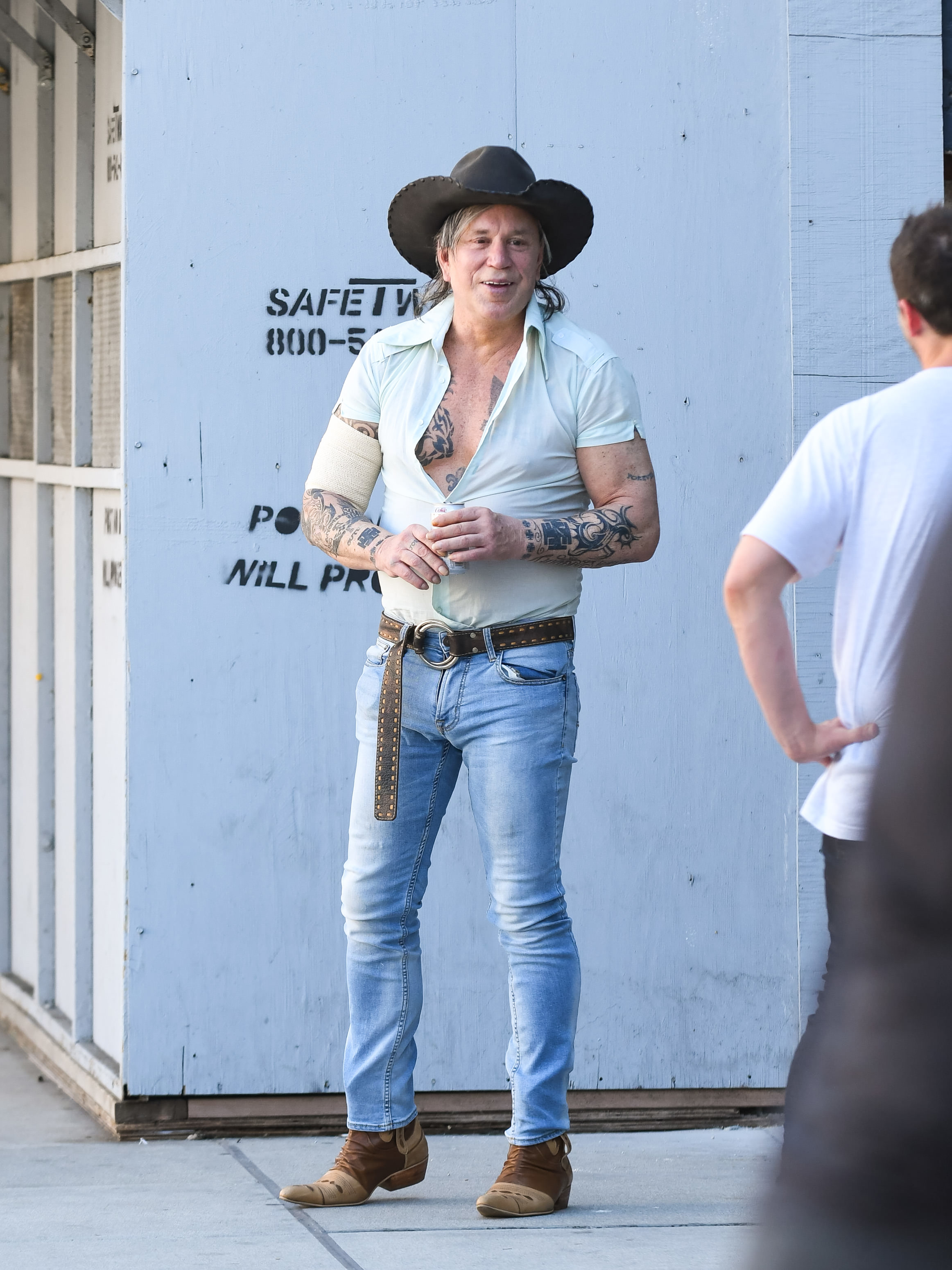 Mickey Rourke in a cowboy hat, light shirt, jeans, and boots, standing outdoors and interacting with another person. Tattoos are visible on arms