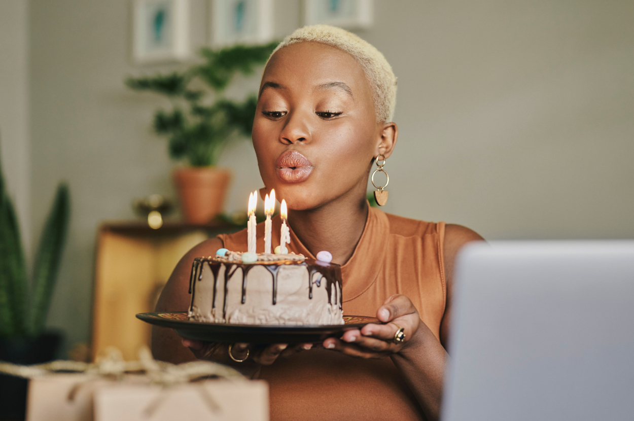 Person blowing out candles on a cake while holding it, celebrating a birthday in a cozy setting with gift boxes nearby