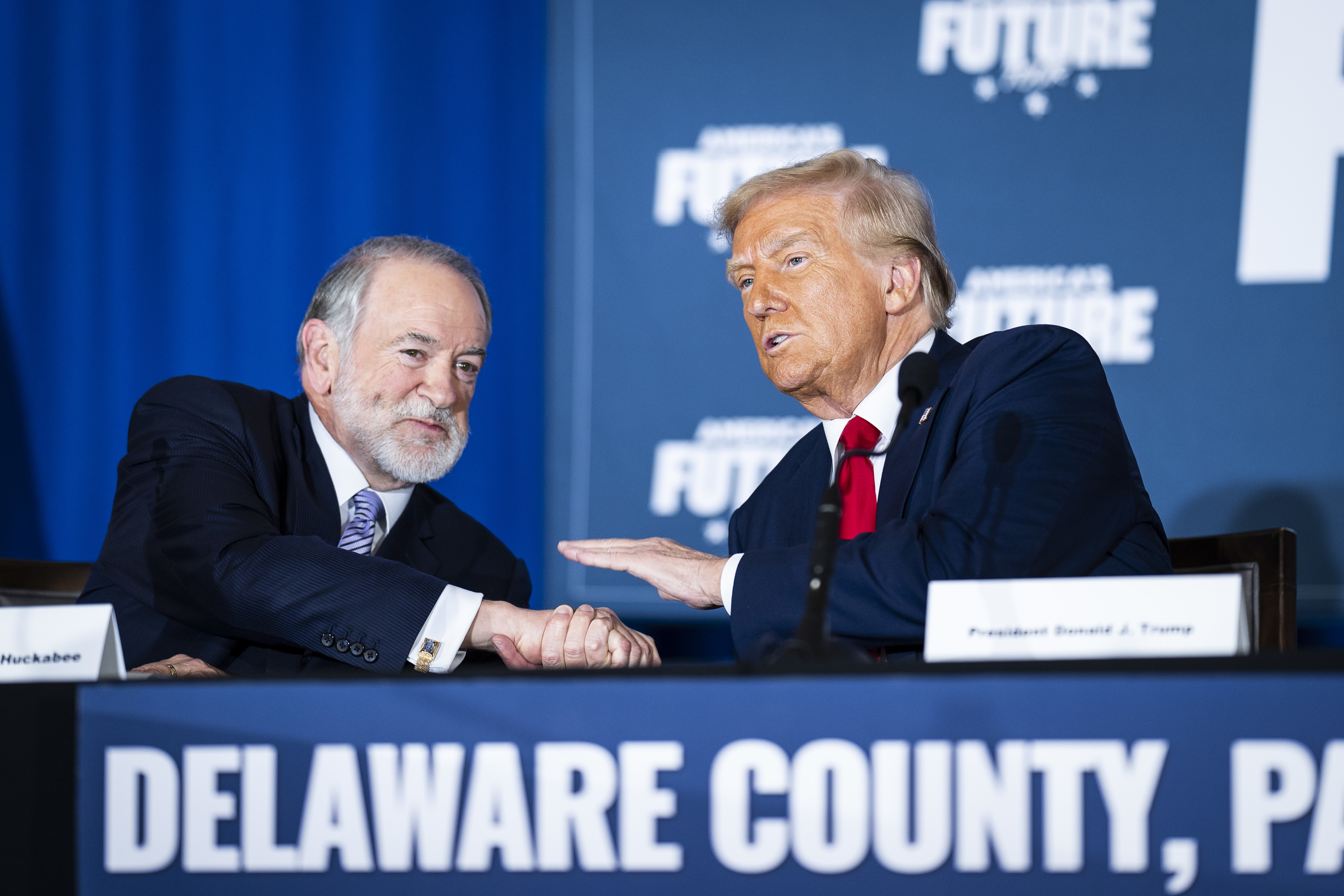 Two men at a panel in Delaware County, PA, shaking hands. One has a suit and tie, the other in a suit gestures while speaking