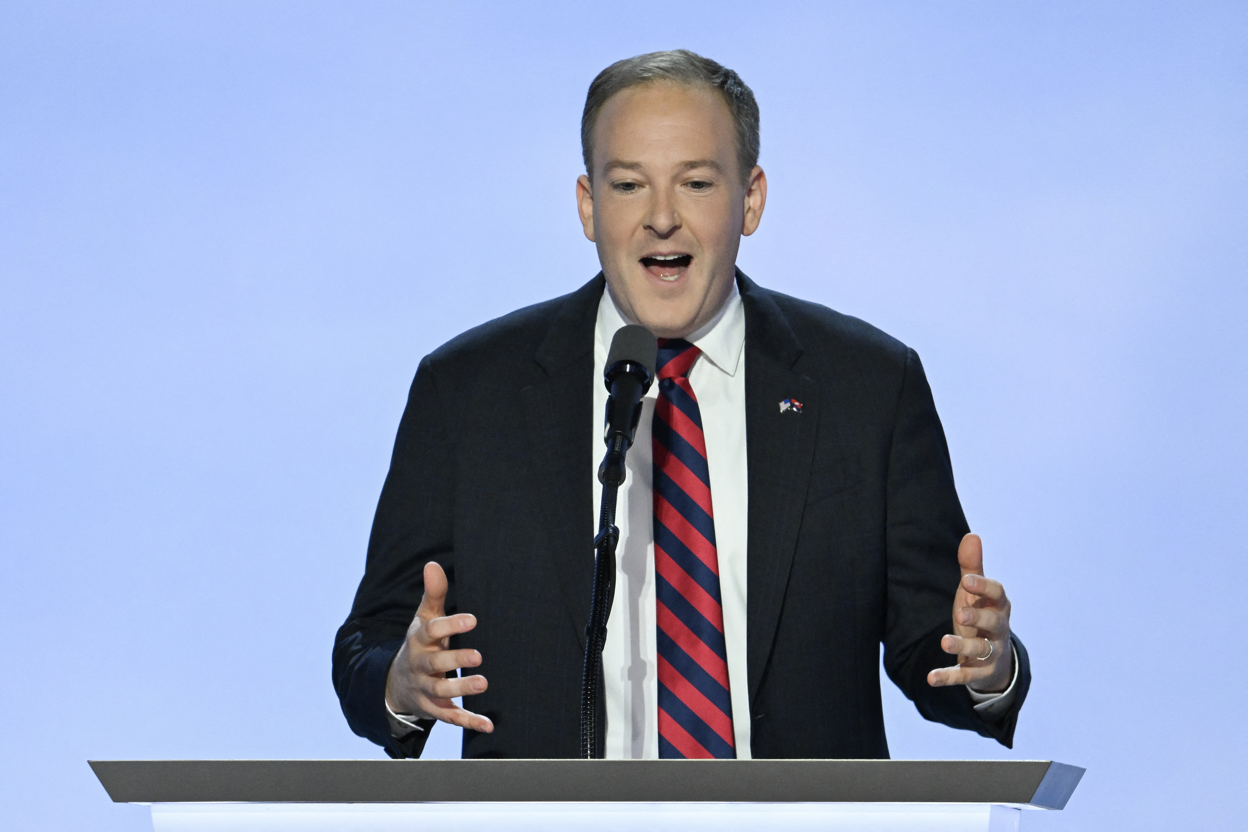 A man speaks animatedly at a podium, wearing a suit with a striped tie against a plain backdrop