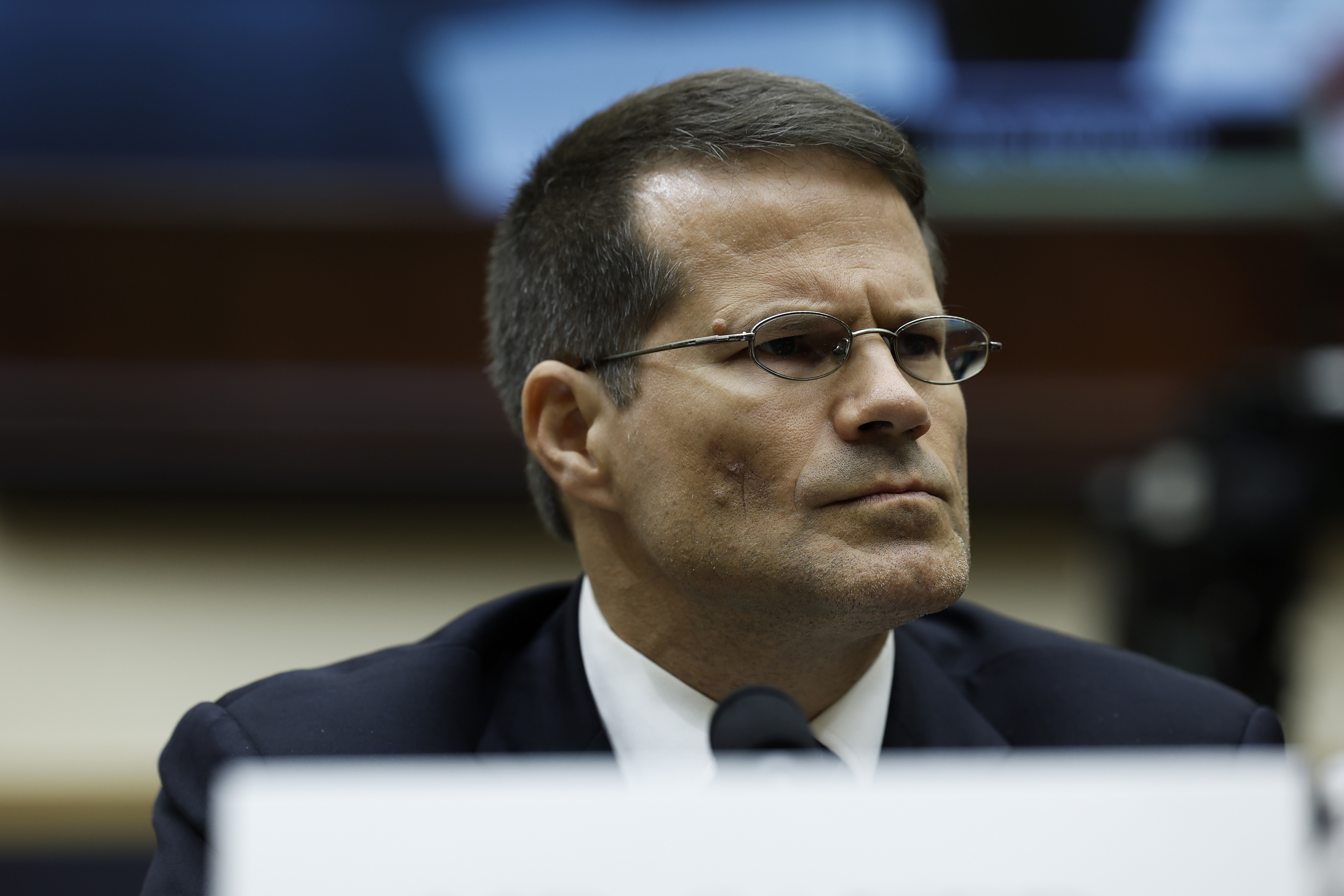 A man in glasses and a suit appears serious at a formal event or hearing, with a blurred background