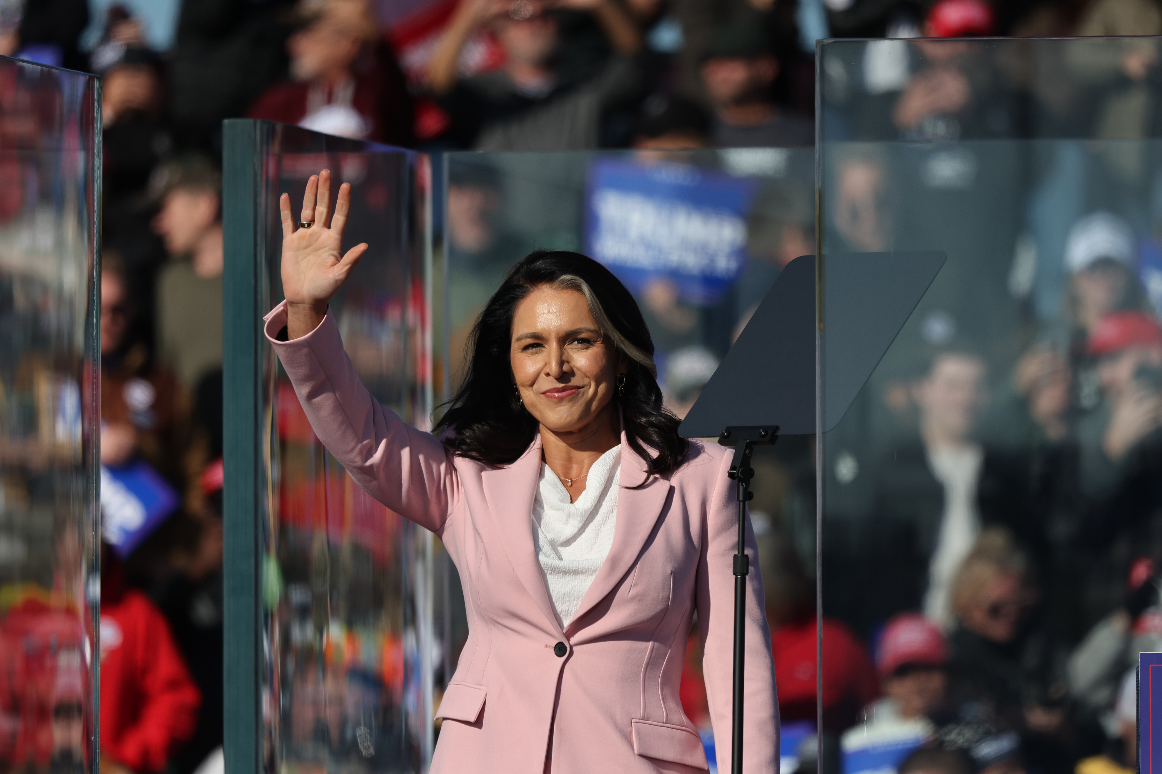 Person in a light suit waves while standing behind a podium during a public event with a crowd in the background