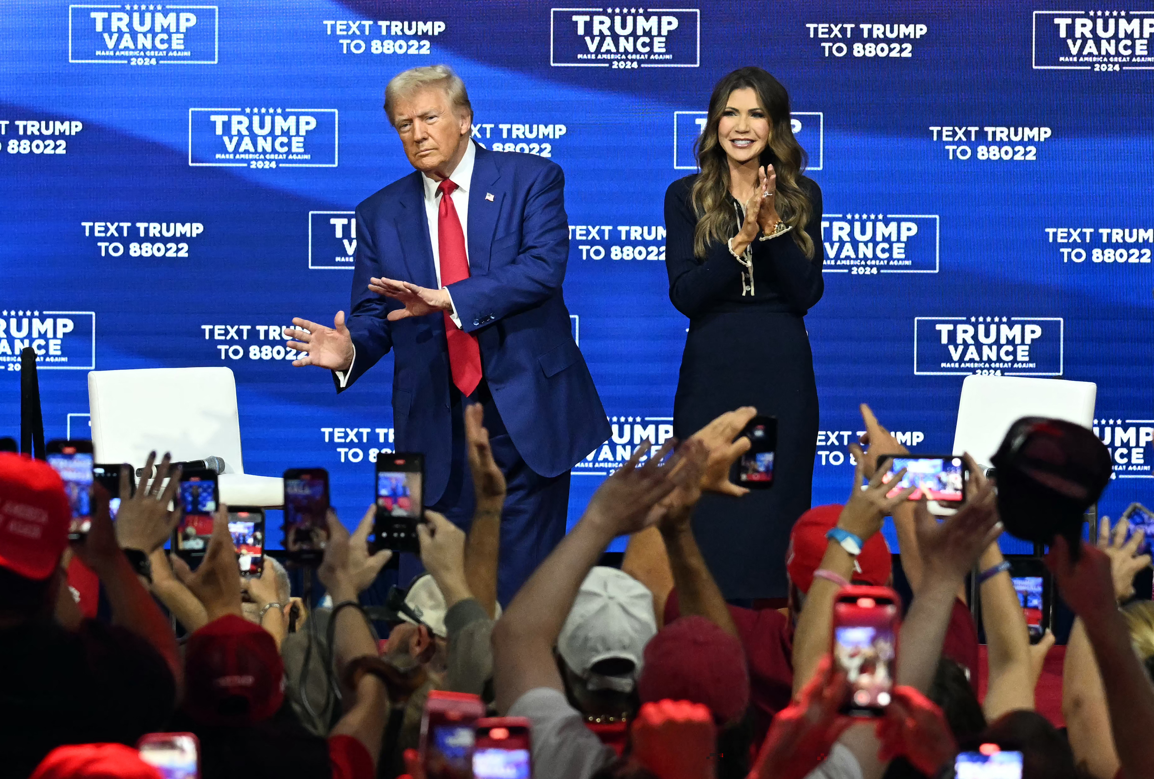 Two people on stage surrounded by a crowd taking photos at a public event. They appear to be addressing the audience