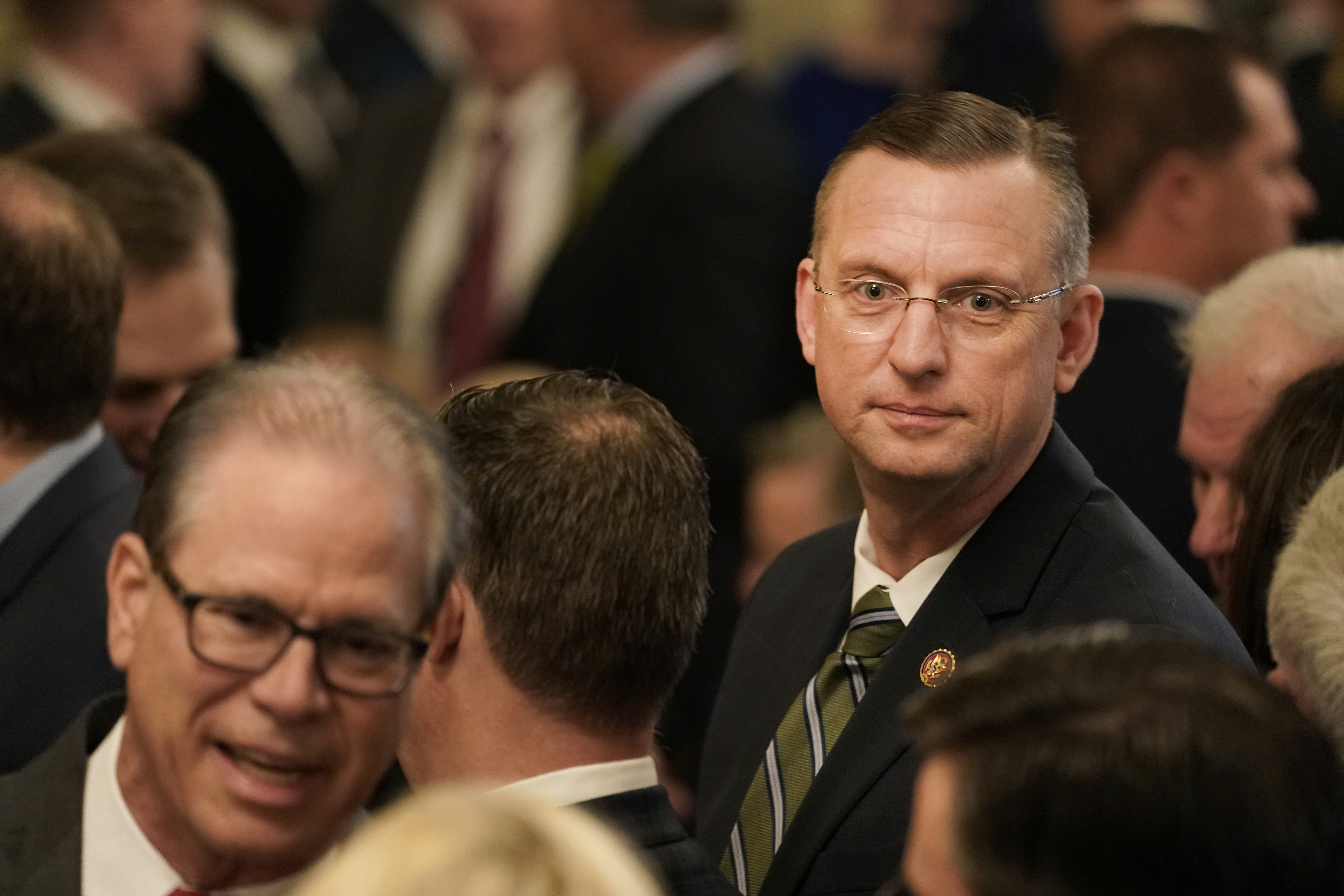 A man in a suit with glasses appears thoughtful amidst a crowd at a formal event