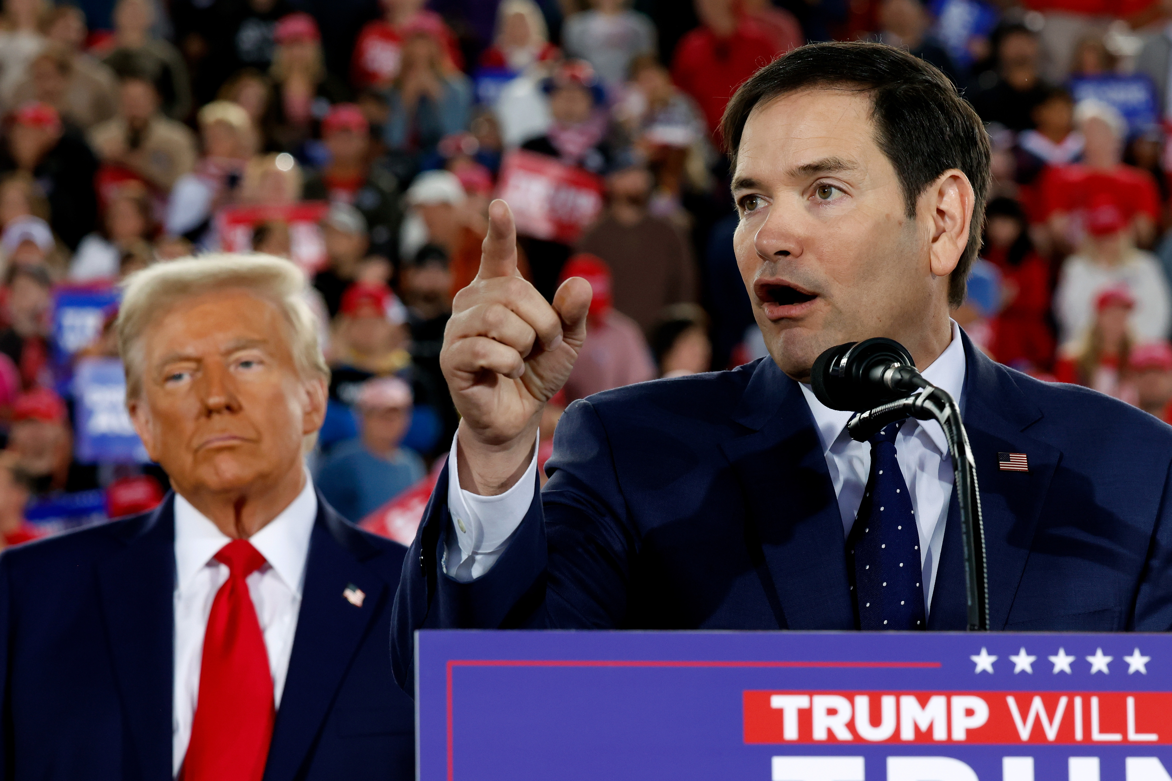 A speaker gestures at a podium during a political rally, with supporters in the background. Another person stands nearby, listening attentively