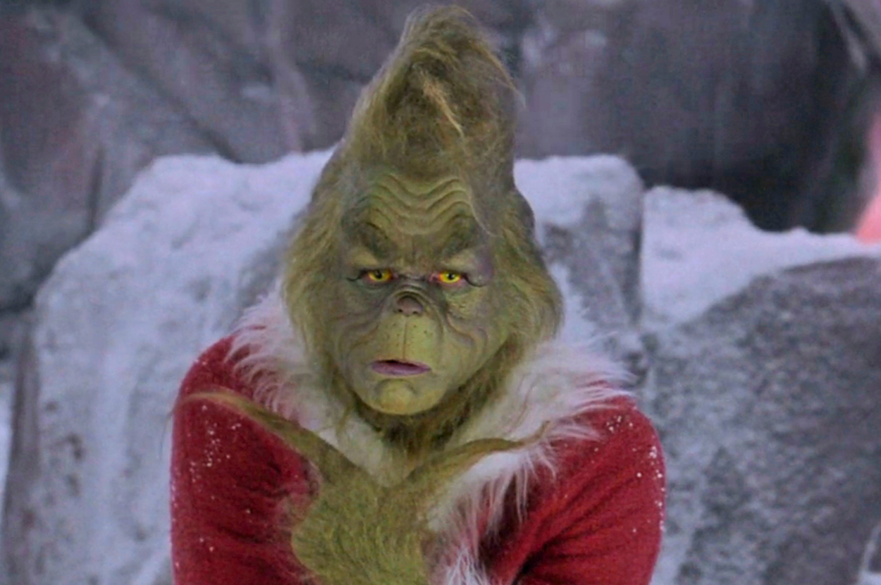 The Grinch, with a puzzled expression, wearing a Santa suit, stands in front of rocky, snowy background