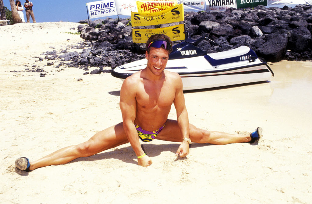 A man does a split on a sandy beach next to a jet ski, smiling, in beachwear and sunglasses