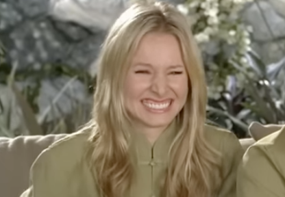 A person with long hair laughing joyfully, sitting on a couch, wearing a casual outfit, with a blurred background