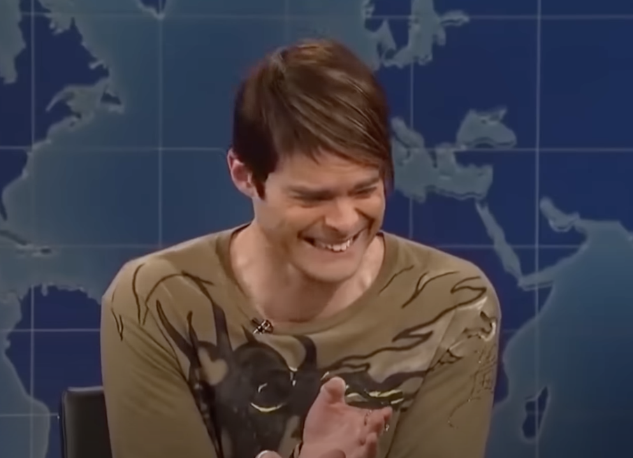 Person laughing on a TV set with a world map backdrop, wearing a printed long-sleeve shirt