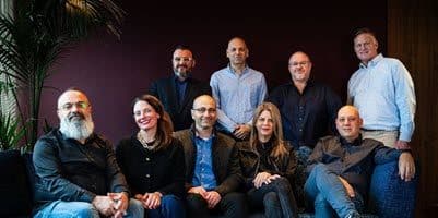 A group of nine people, both men and women, are seated and standing together indoors, posing for a photo. They are dressed in casual to business attire. A plant is visible to the left, set against dark purple and wood tones.