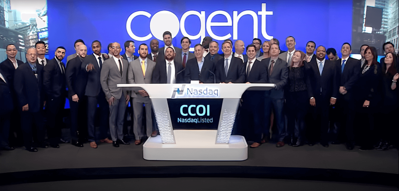 A large group of people stands on a stage in front of a Nasdaq podium with Cogent Communications displayed on a screen behind them. The diverse group, clad in business attire, smiles brightly as they pose for a photograph.