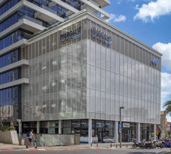 A modern glass and steel building with the words Margalit Startup City on the facade rises in Tel Aviv. It has multiple stories set against a clear sky. A person walks on the sidewalk next to bicycles parked nearby, capturing the city's bustling innovation hub.