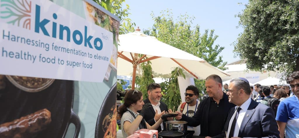 People are gathered at an outdoor food event. A booth with a kinoko banner showcases their products. Among the attendees a man in sunglasses engages with presenters. Umbrellas and trees provide shade in the sunny setting.