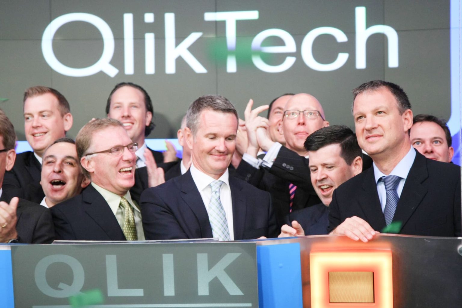 A group of men in suits celebrates at a stock exchange event. They stand in front of large digital displays showing the QlikTech logo. Some are clapping, and others look excited, marked by confetti falling around them.