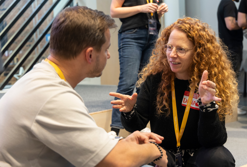 A woman wearing a black t-shirt is talking to a man sitting in-front.