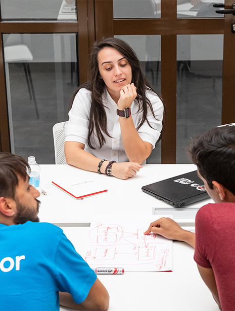 Woman is speaking to two men around a table. One man with his back to the camera is wearing a t-shirt that says "mentor".