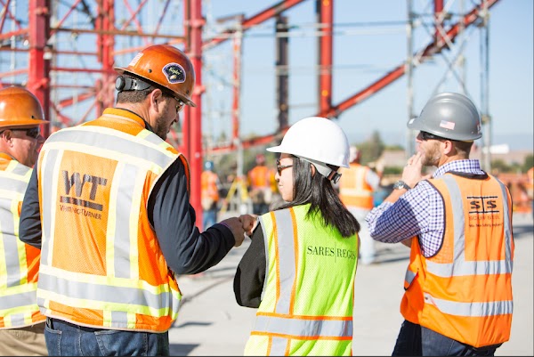 Four people wearing construction gear talking as one of the “smile truss” pieces is lifted into place.