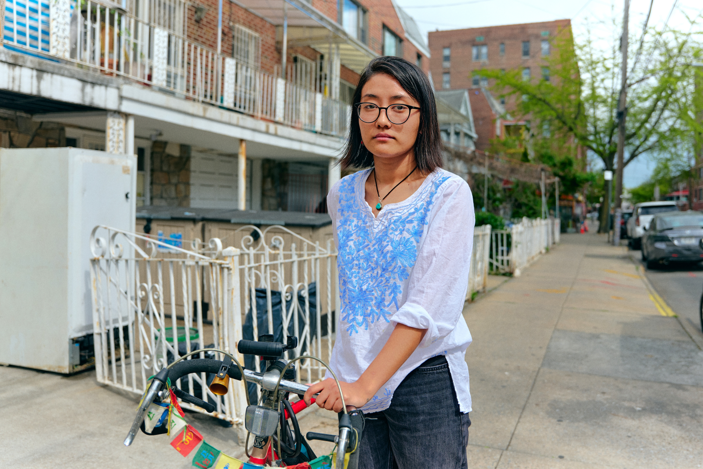 A young Asian woman with glasses and short black hair standing with her bike on the sidewalk.