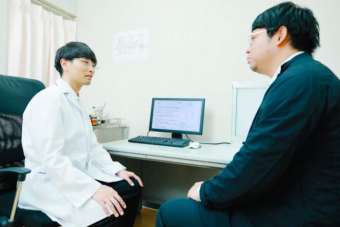 Man in a lab coat in front of a computer sits talking to another man