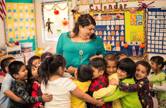 Smiling woman in a school classroom surrounded by a group of young children.