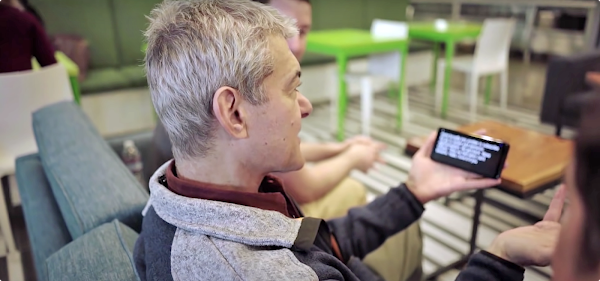 Google research scientist Dimitri Kanevsky sits on a blue couch conversing with colleagues. From behind his right shoulder, his left hand is seen holding a phone with text-to-speech captions visible on the screen.