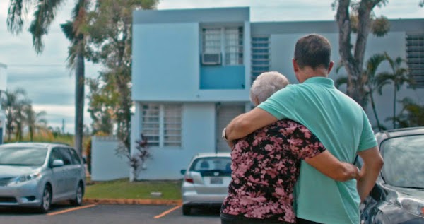 A woman with short gray hair wearing a black and pink shirt and a man with dark short hair wearing a light blue shirt embrace in a parking lot as they look past blue buildings and palm trees to the horizon.