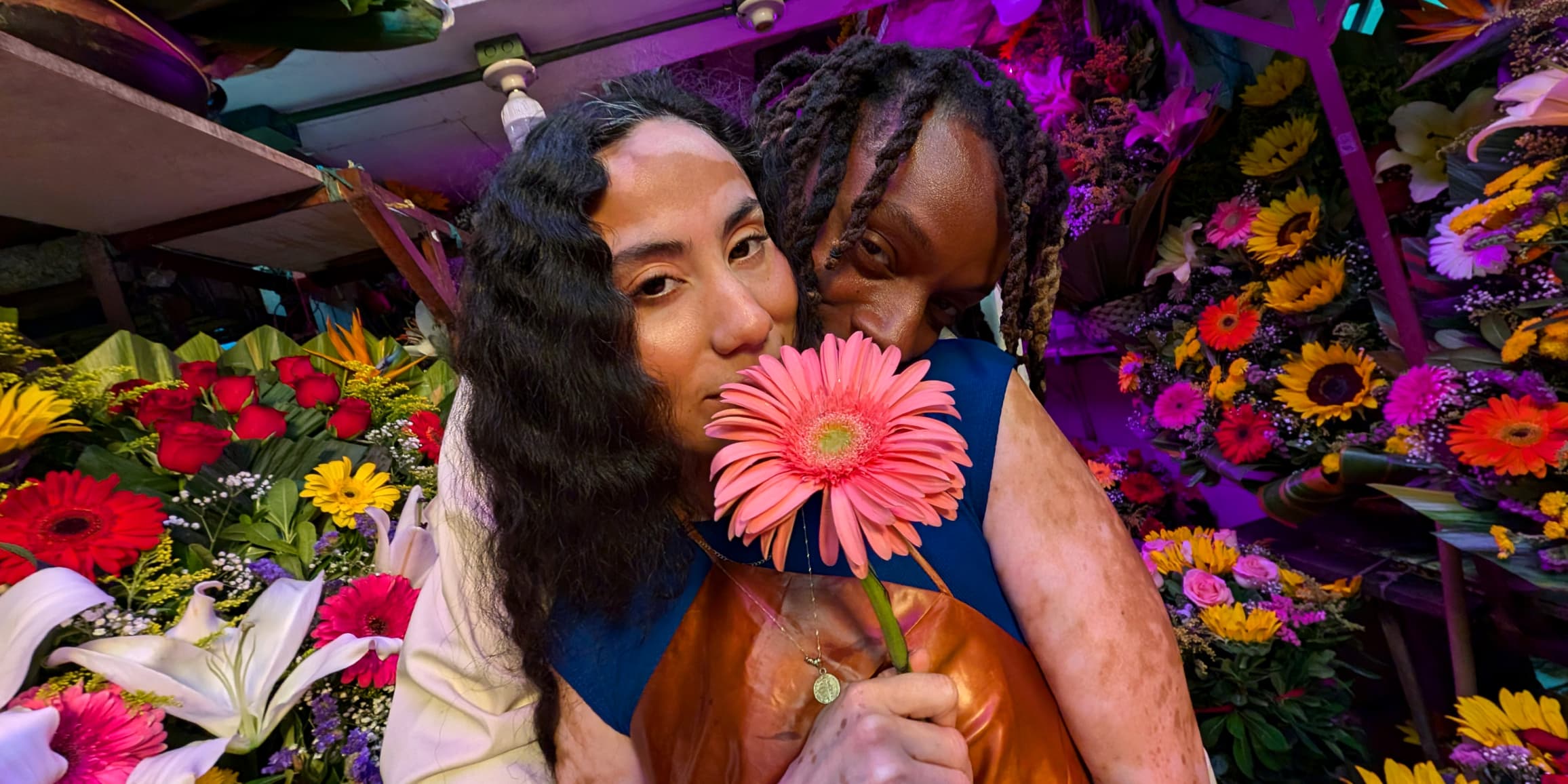 Two people pose facing the Pixel Camera, surrounded by flowers. Colours and skin tones are vividly captured by Pixel 9.