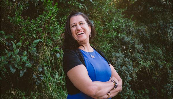 Mor Schlesinger stands smiling in front of a green background of plants and trees. She has medium-length dark brown hair and is wearing a blue top and black sweater.