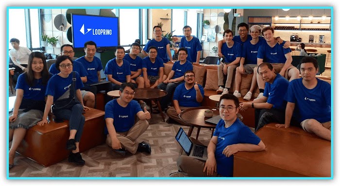 Group of people in blue Loopring tee shirts gathered in front of a monitor displaying Loopring logo.