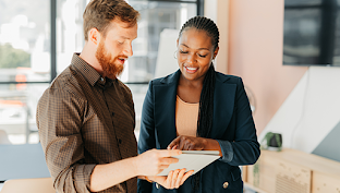 A white man in a brown button-up reviewing a project with a Black female coworker wearing a blue suit in a conference room.