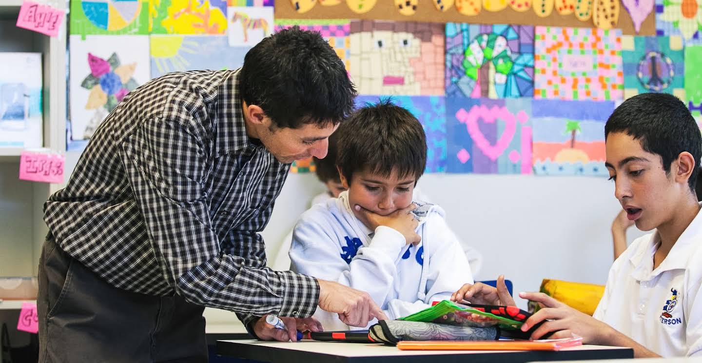 A male teacher in a checkered black and white shirt helping a student at their desk.