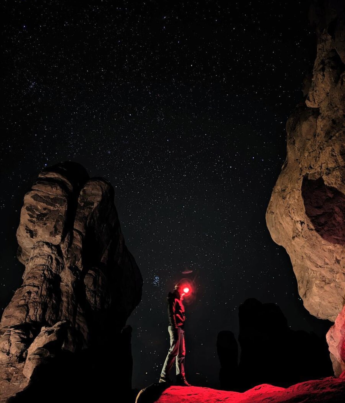 A person stands in the dark besides two columns of rock, wearing a red light on their head. The night sky is speckled with stars.