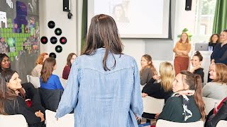 Woman standing and talking to a group of women, her back facing the camera.