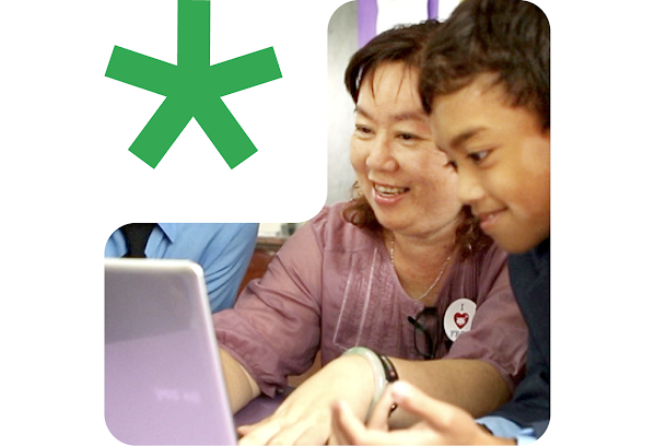 A smiling teacher types on a computer with a young student next to her.