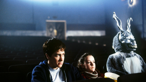 A screenshot from Donnie Darko of Jake Gyllenhall sitting in a movie theater next to a man in a creepy rabbit suit