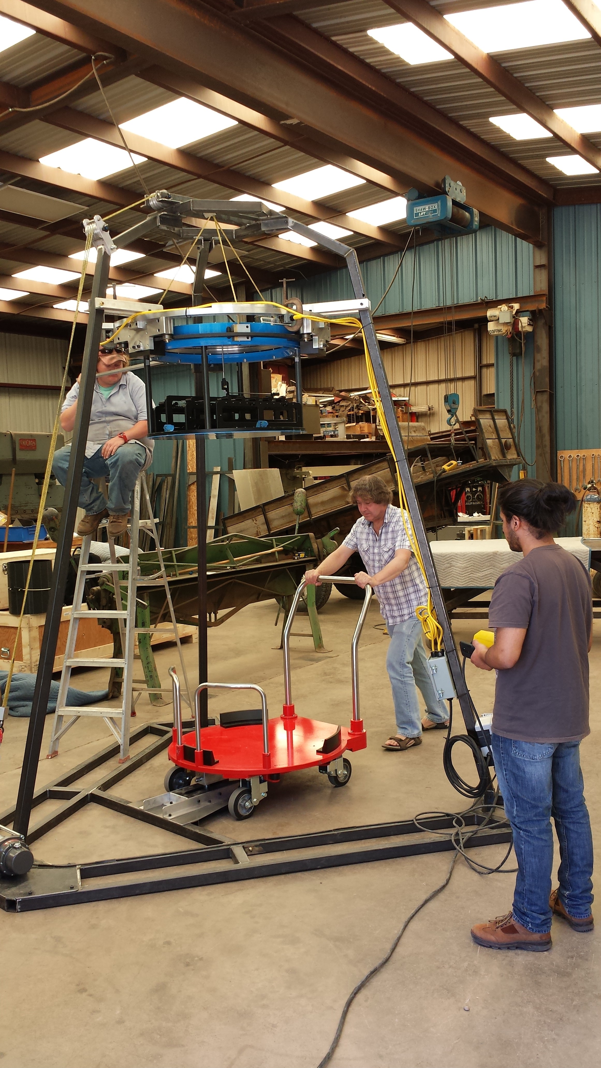 Christian and Jamie swapping out a plug plate cartridge with the Mock Up and Training Facility tripod (the big steel frame), cartridge (the blue object suspended from the tripod) and dolly, which will be used to transport plug plates to and from the telescope. Christian y Jaime intercambiando el cartucho de la placa conexión con la maqueta y el trípode de capacitación (la estructura de acero grande), el cartucho (el objeto azul suspendido del trípode) y el carro, que será utilizado para transportar las placas de conexión hacia y desde el telescopio.